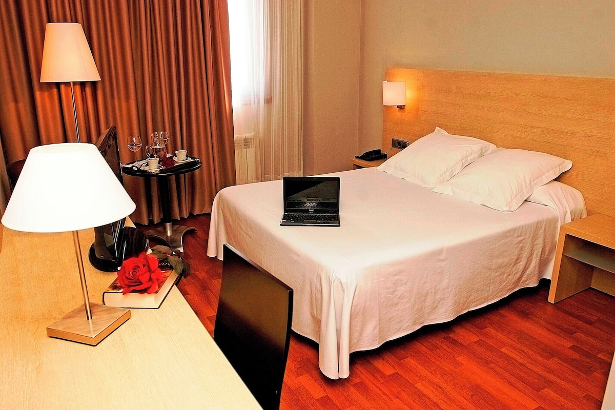 A hotel room with a bed, a desk with a lamp, a laptop, and a bedside table with a phone and a tray of items.