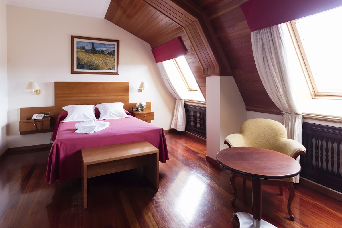 A hotel room with a wooden bed, a bench, a round table, a chair, a nightstand, and a painting on the wall.