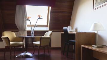 A room with a wooden ceiling, a desk with a lamp, two chairs, a small table with a vase, and a window with curtains.