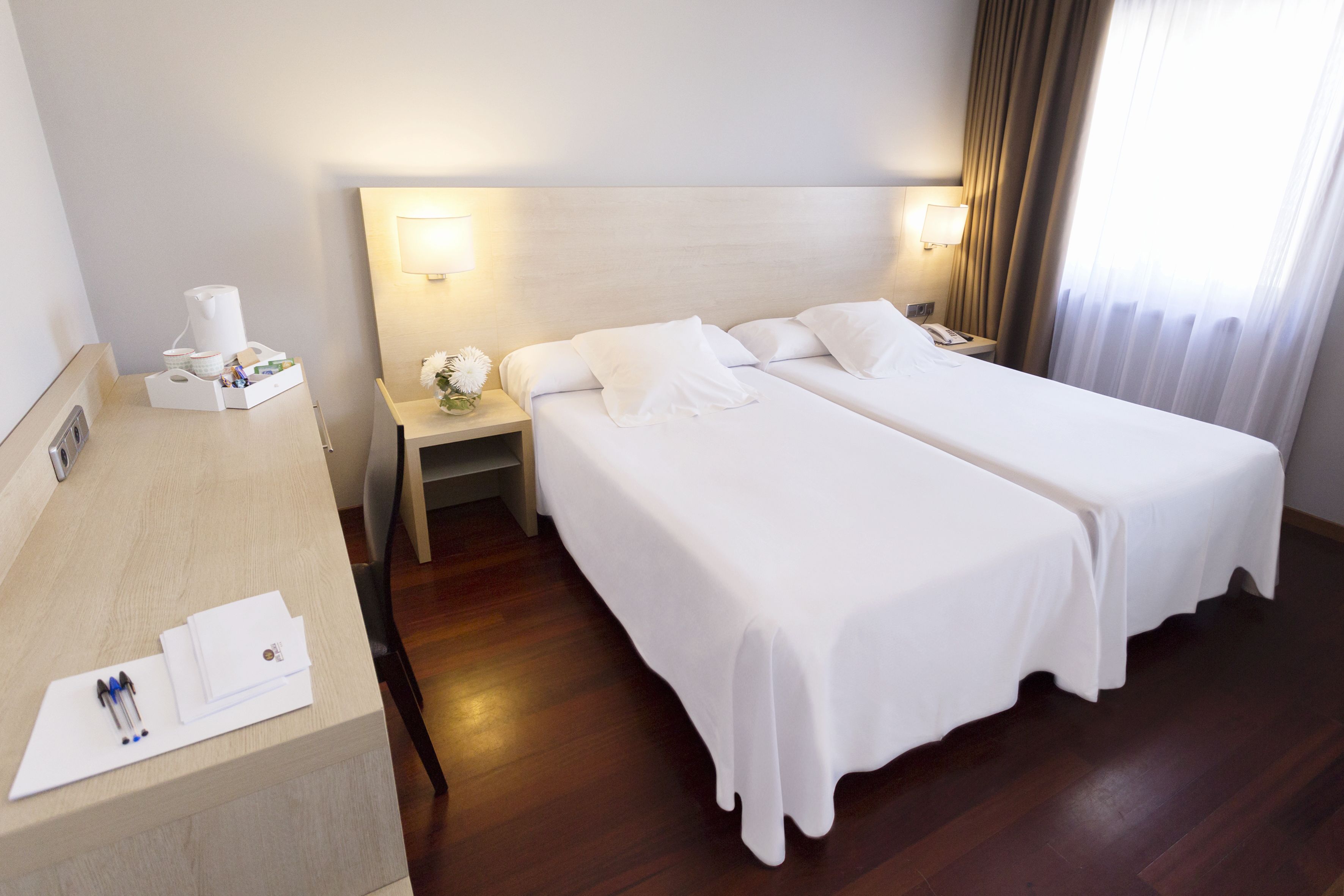 A hotel room with two single beds, a wooden desk with a coffee maker, and a nightstand with a lamp.