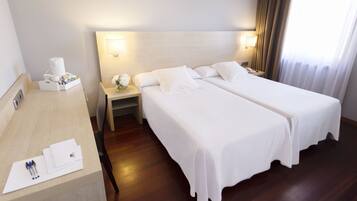 A hotel room with two single beds, a wooden desk with a coffee maker, and a nightstand with a lamp.