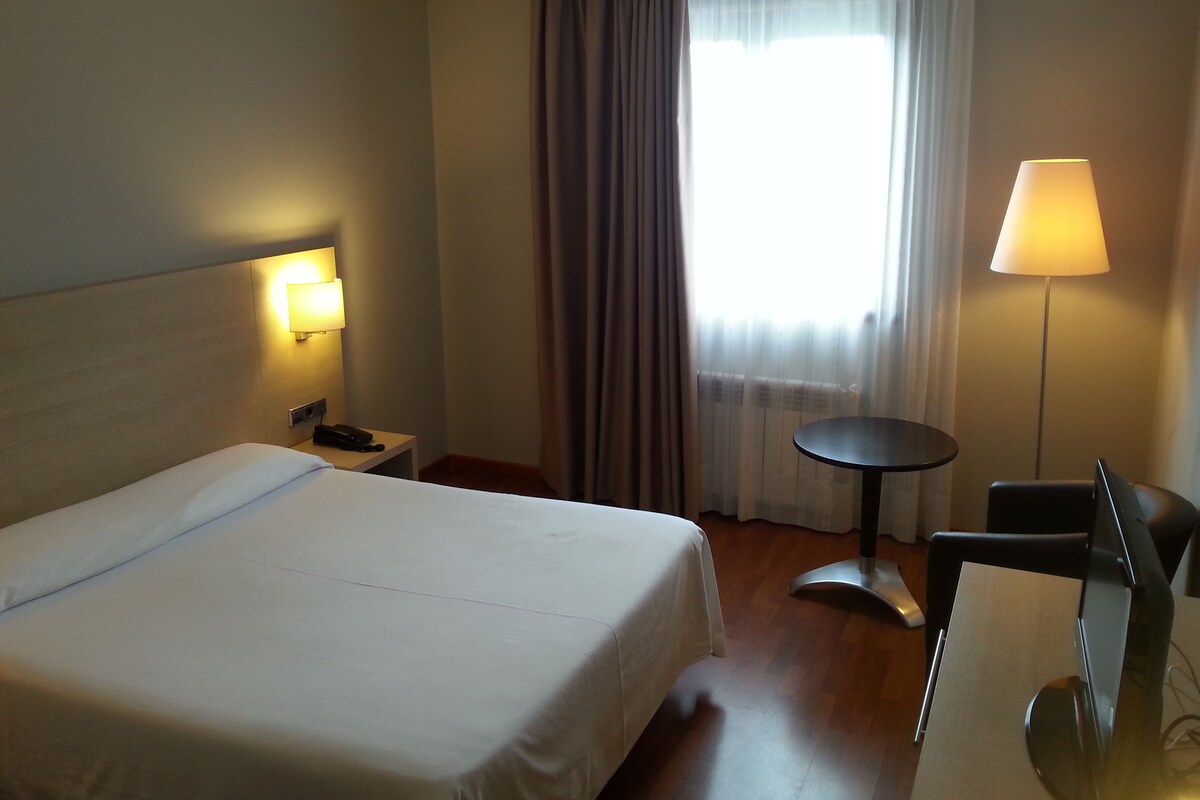 A hotel room with a bed, bedside table, lamp, chair, and a view of the outdoors through a window with curtains.