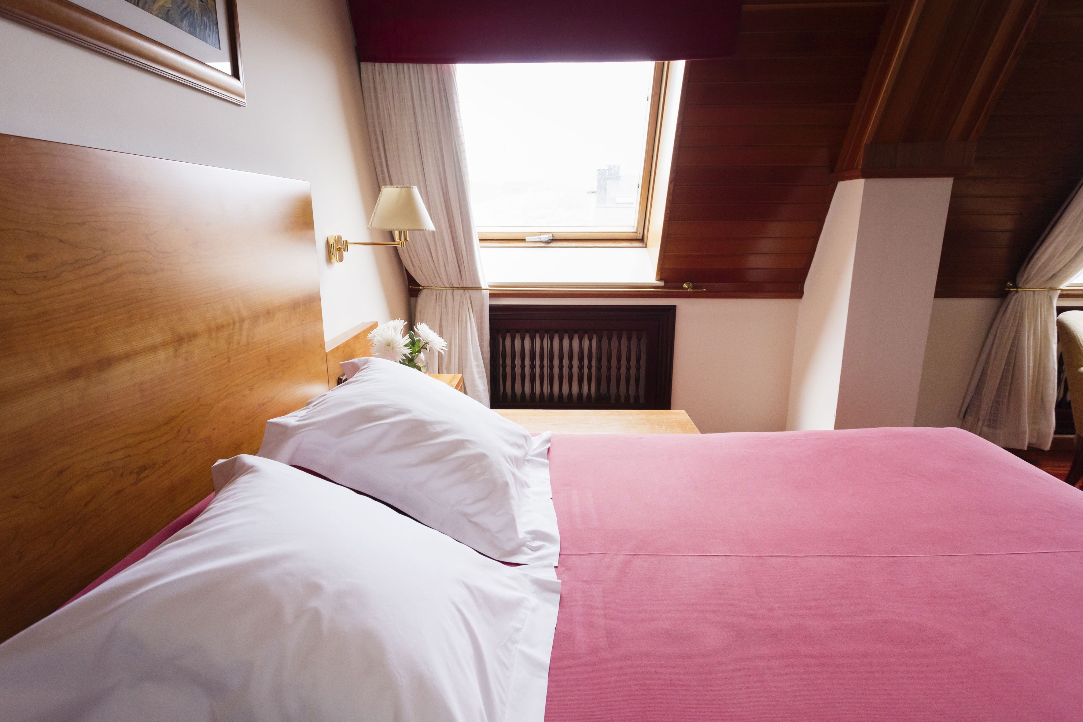 A hotel room with a wooden headboard, a pink bedspread, white pillows, a nightstand with a lamp, and a window with curtains.