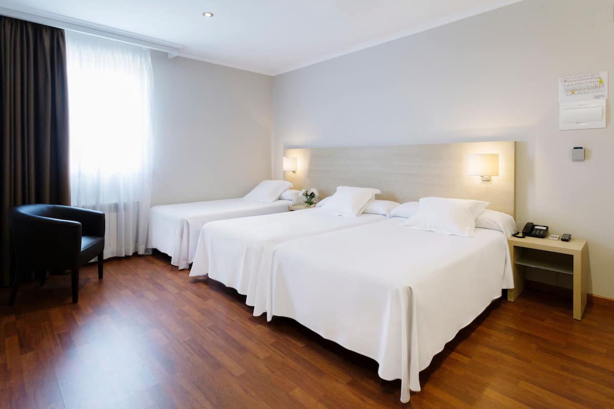 A hotel room with two beds, a chair, a bedside table with a lamp, and a wall clock.