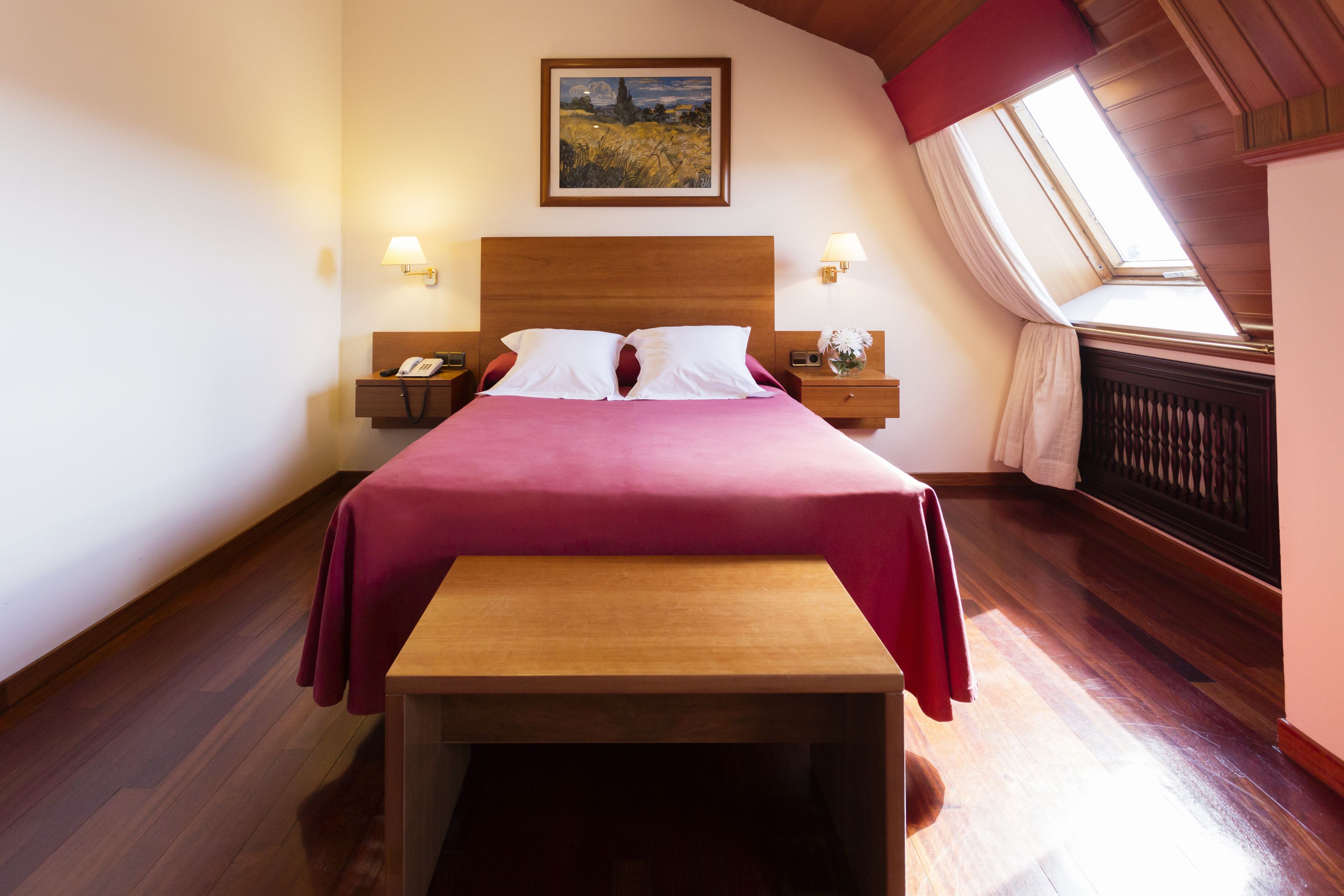 A neatly arranged bedroom with a large bed, bedside lamps, a nightstand, a wooden bench, and a skylight.
