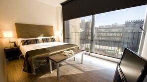 Habitación de hotel con cama, lámparas de noche, una mesita con una bandeja de objetos, televisor y un ventanal con vistas a la ciudad.