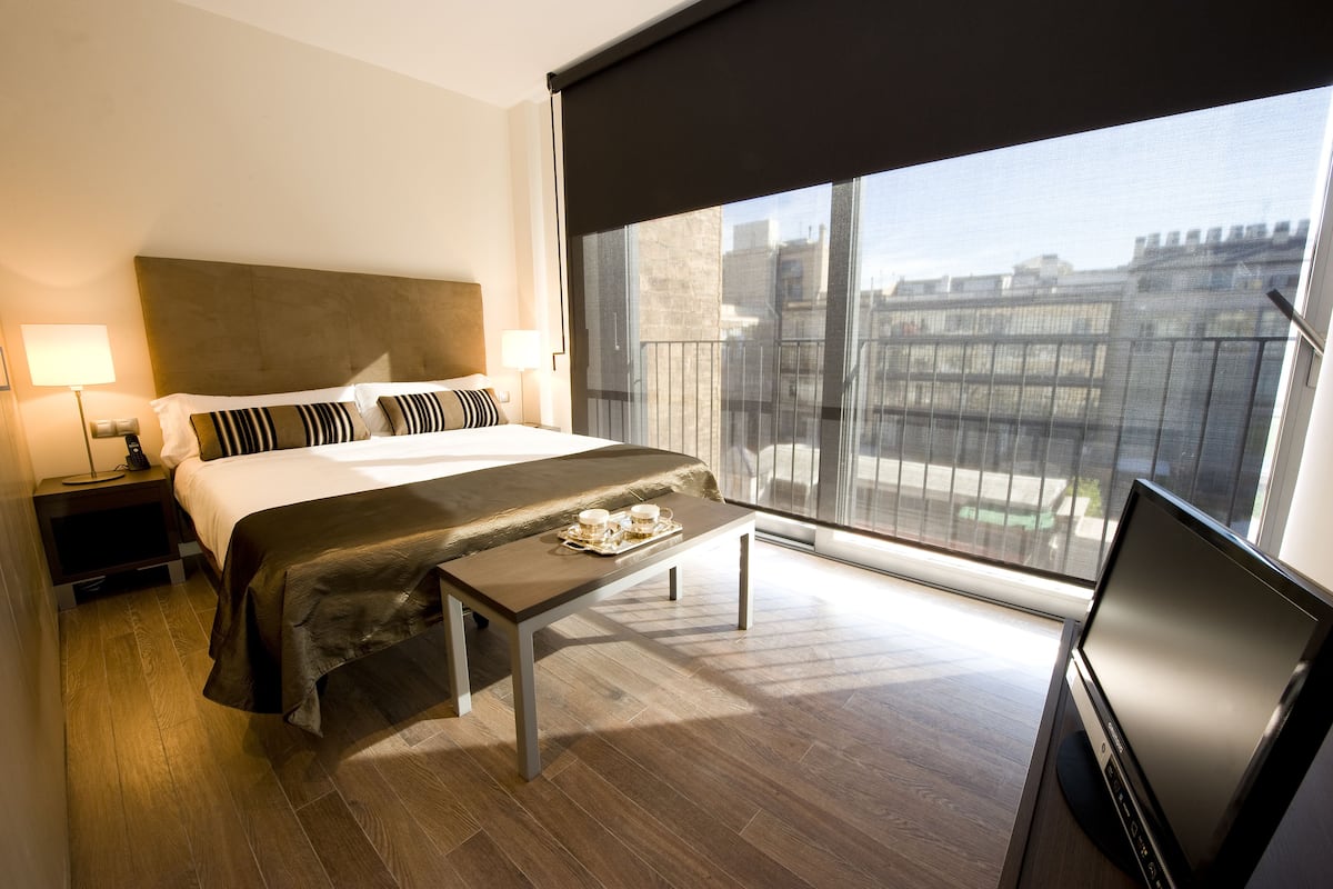 A hotel room with a bed, bedside lamps, a small table with a tray of items, a TV, and a large window offering a city view.
