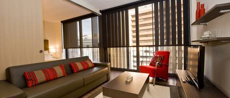 A modern living room with a sofa, a coffee table, a red armchair, a TV, and a large window with blinds.