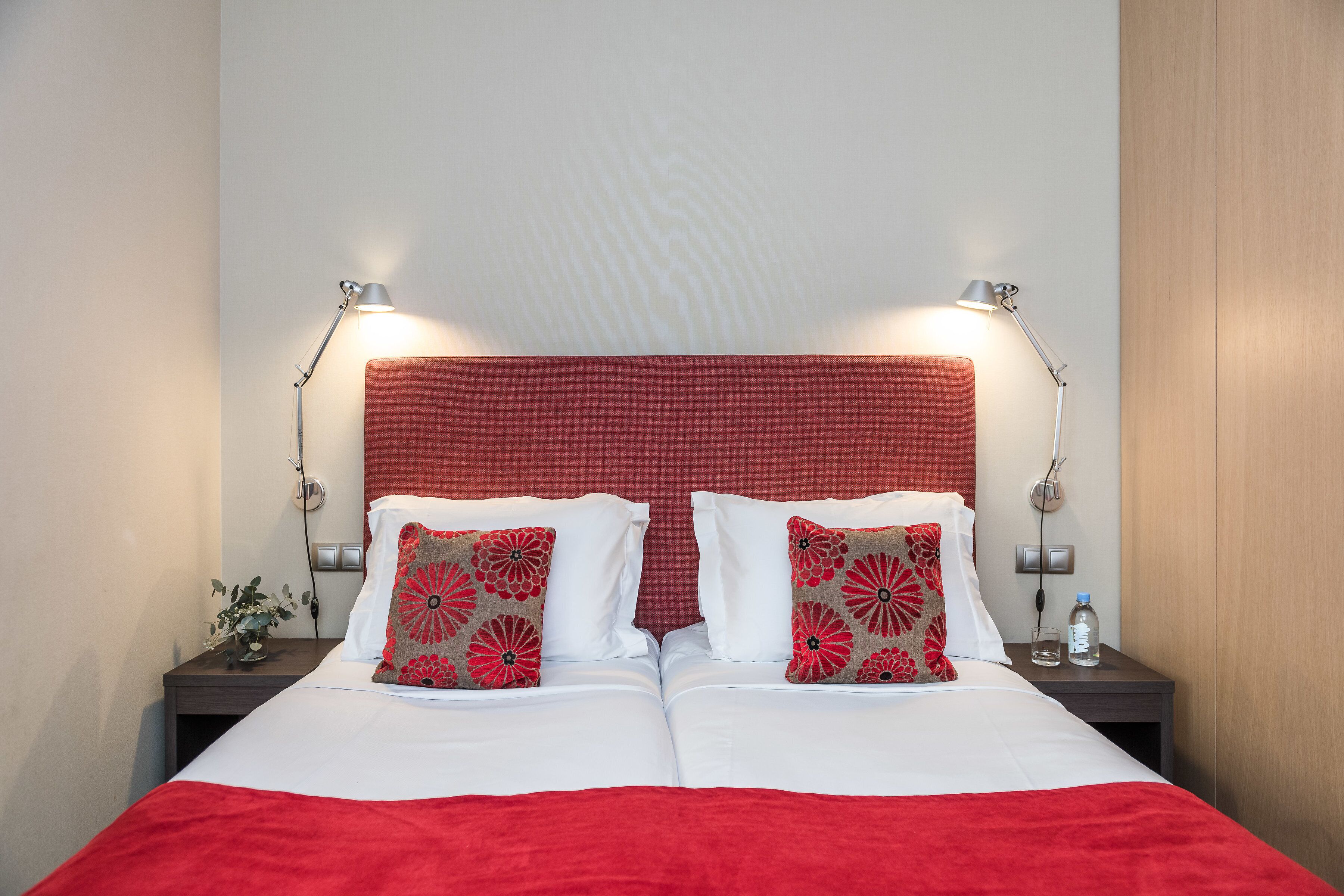 A neatly made double bed with white linens and red accents, two bedside lamps, and a small table with a water bottle and a glass.