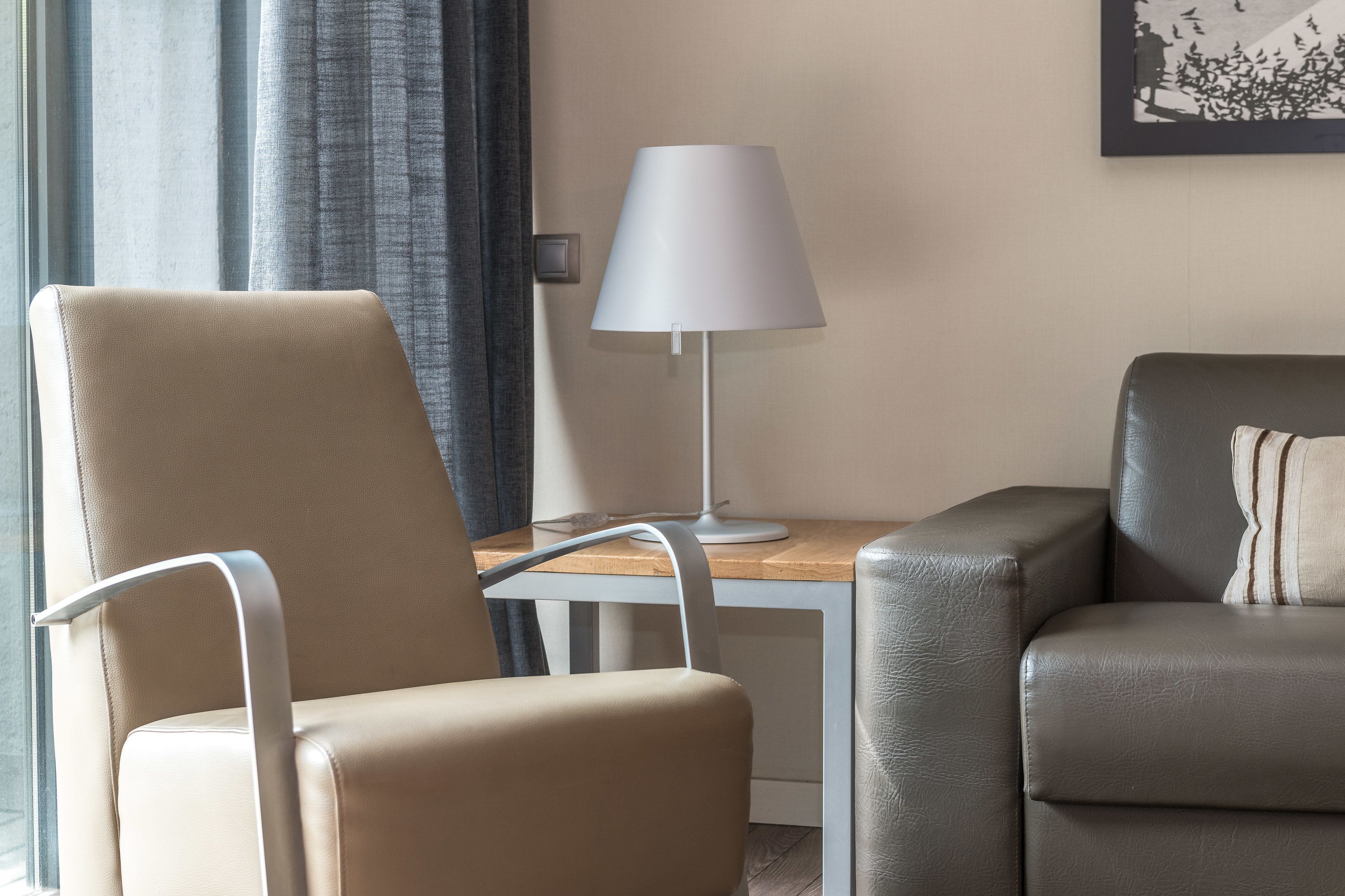 Une chambre d’hôtel moderne avec un fauteuil beige, un canapé en cuir foncé, une lampe blanche et une table en bois.