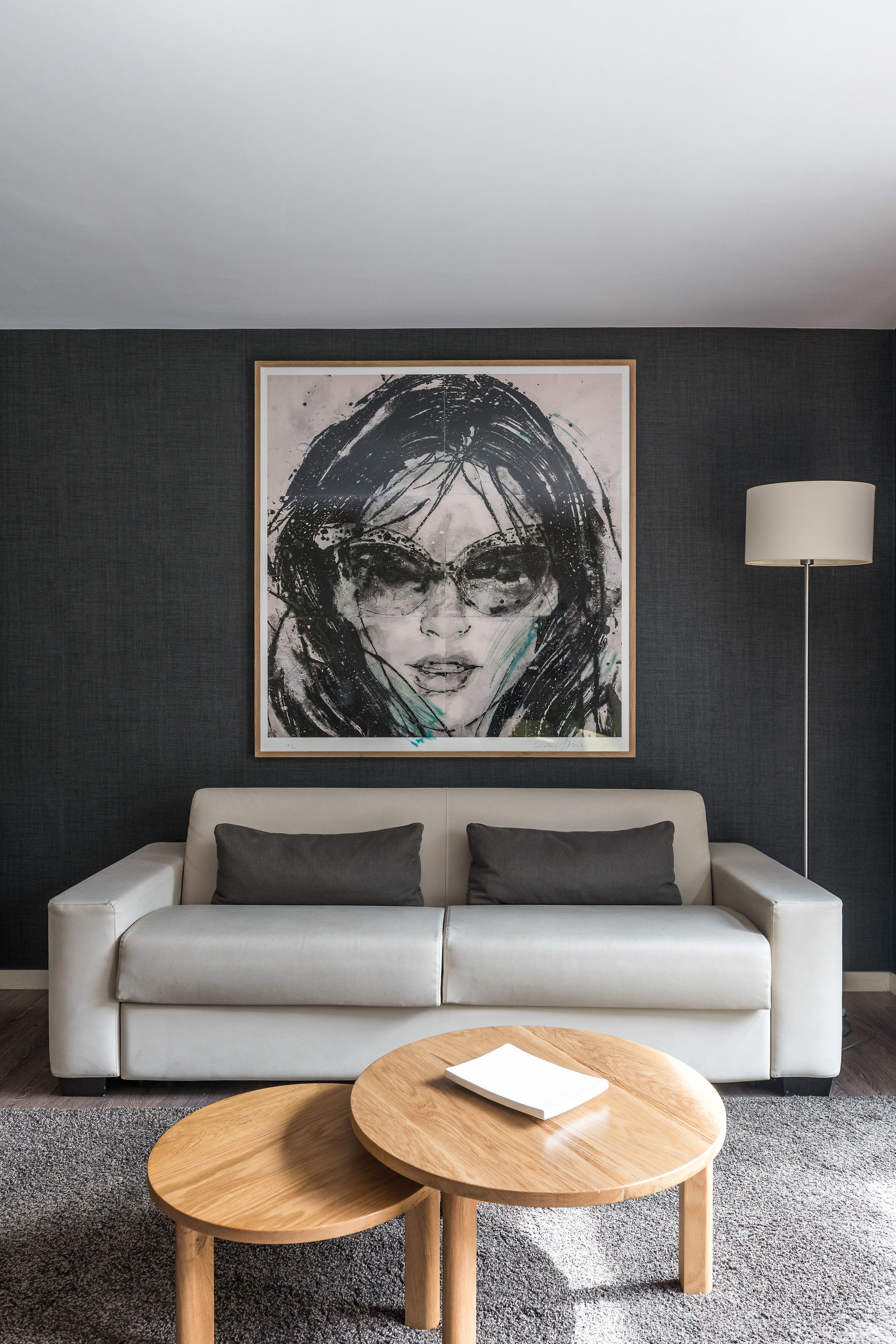 A modern living room with a grey sofa, two round wooden coffee tables, a large abstract painting on the wall, and a floor lamp.