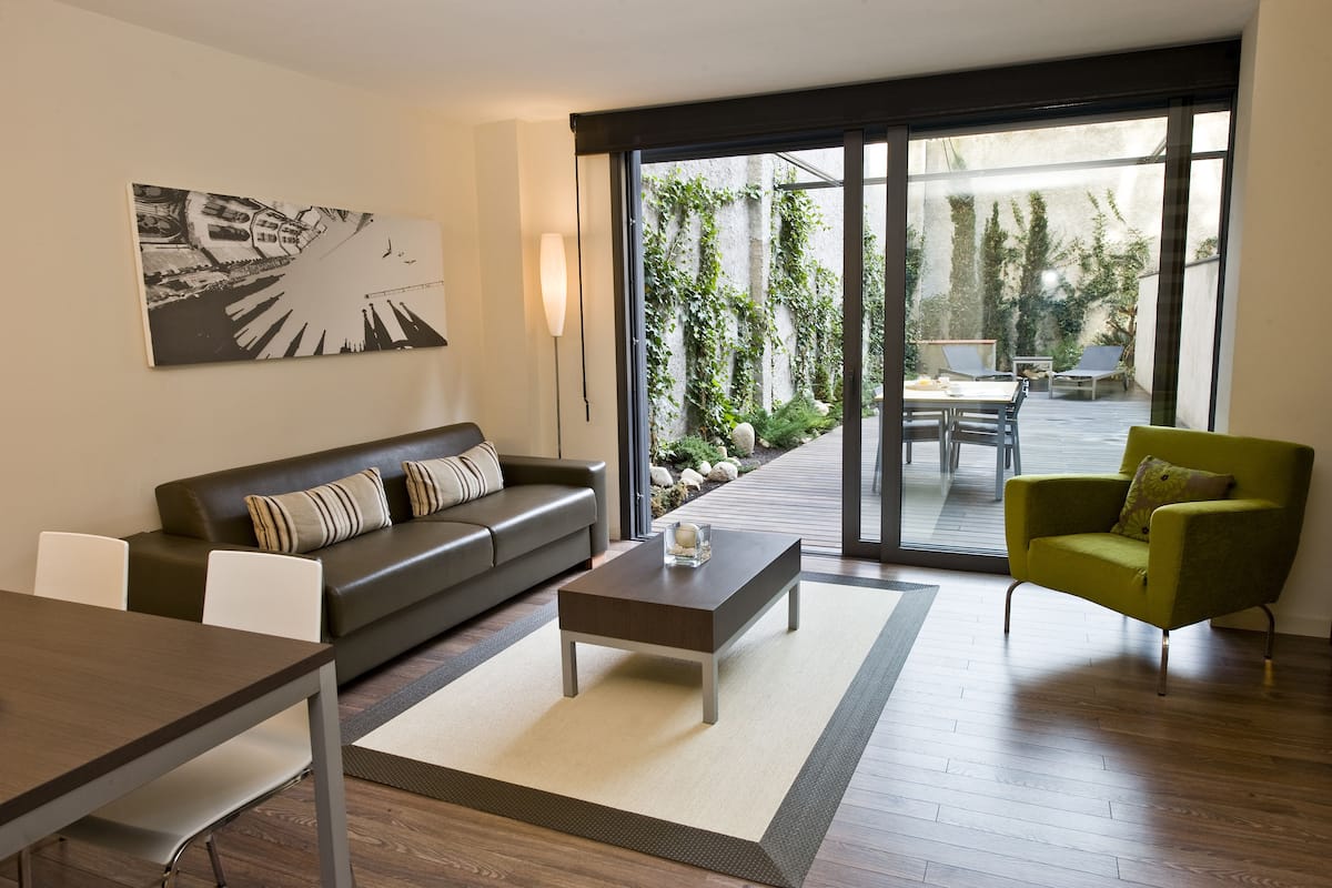 A modern living room with a sofa, coffee table, and armchair, a dining area with chairs, and a large sliding glass door leading to an outdoor patio with a table and chairs.