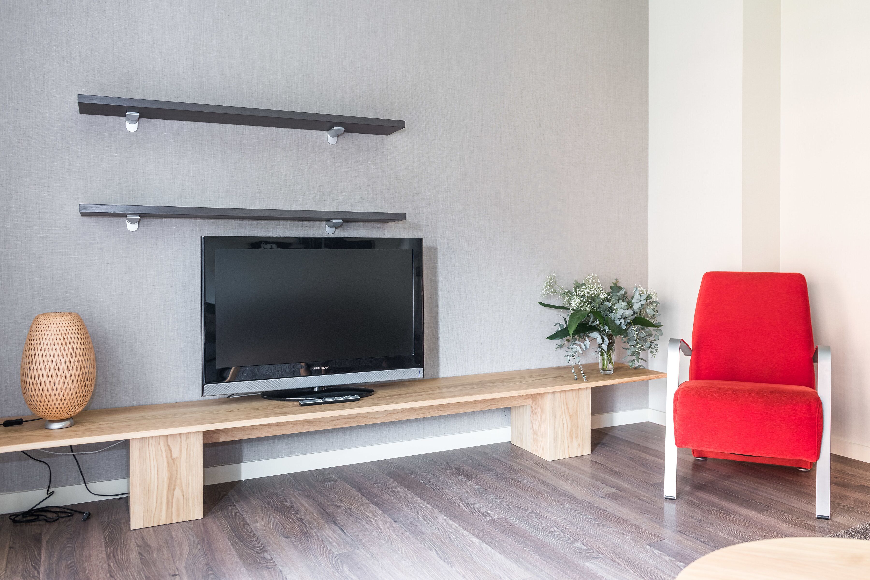Una sala de estar moderna con un televisor de pantalla plana montado en la pared, un banco de madera, una silla roja y un jarrón decorativo.