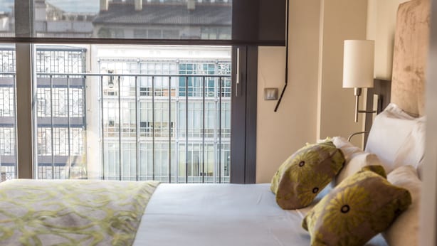 Habitación de hotel con un ventanal grande, una cama con cubrecama estampado, dos almohadones decorativos y una lámpara de noche.
