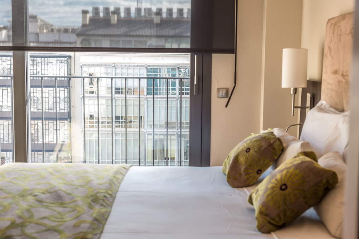A hotel room with a large window, a bed with a patterned bedspread, two decorative pillows, and a bedside lamp.