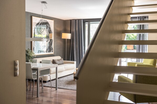 Una sala de estar moderna con un sofá blanco, una mesa de comedor y sillas, un ventanal con cortinas y una escalera al frente.