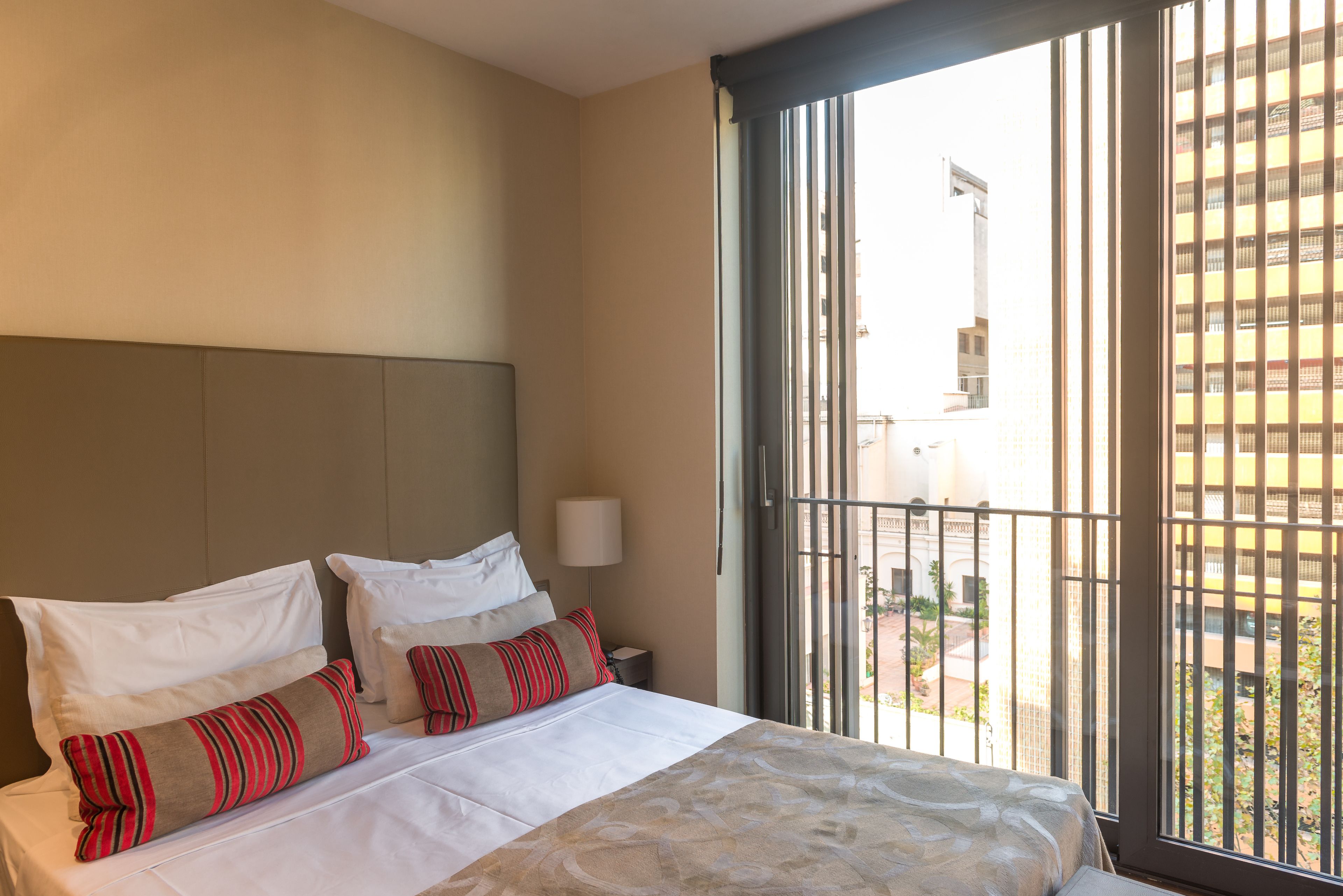 A hotel room with a bed, pillows, a nightstand, a lamp, and a view through a window.