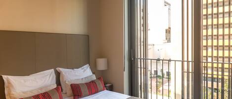 A hotel room with a bed, pillows, a nightstand, a lamp, and a view through a window.