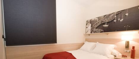 A neatly made bed with a red blanket, a wooden nightstand with a lamp, and a wall-mounted picture of a coastal scene.