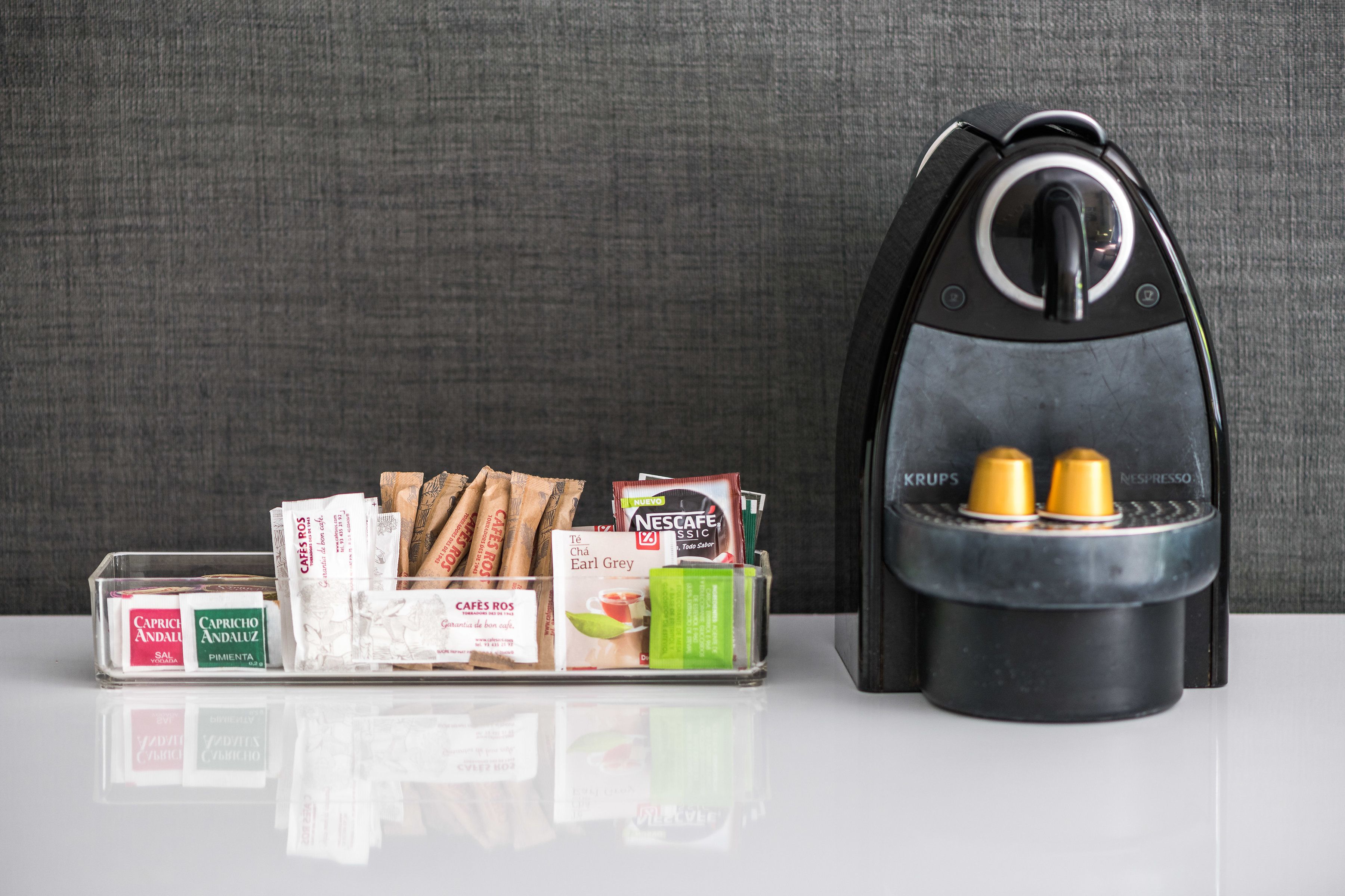 Máquina de café com duas chávenas, porta-cápsulas de café com várias cápsulas e um saquinho de chá.