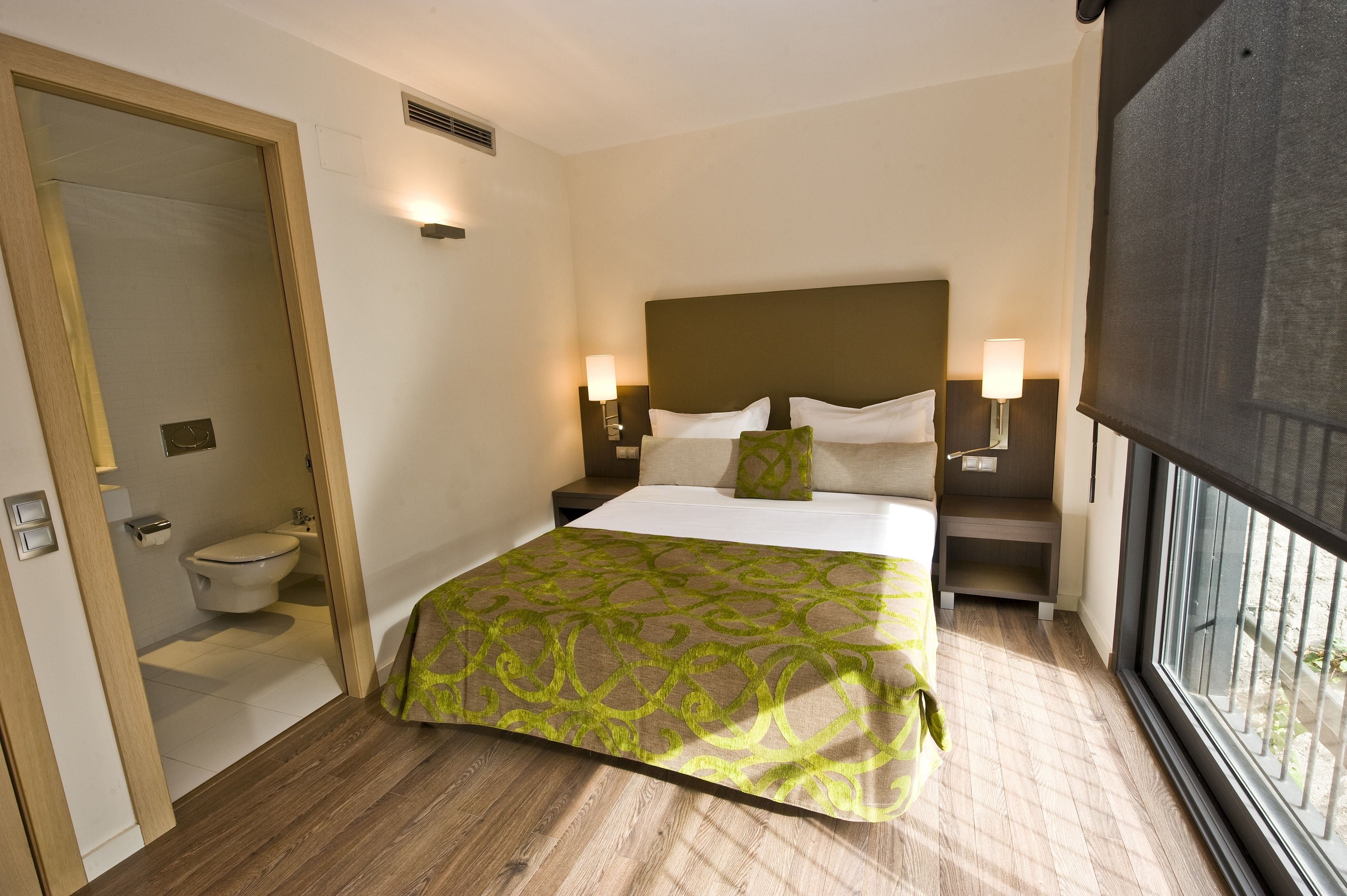 A hotel room with a large bed, two bedside tables with lamps, a window with a view, and a door leading to a bathroom.