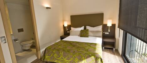 A hotel room with a large bed, two bedside tables with lamps, a window with a view, and a door leading to a bathroom.