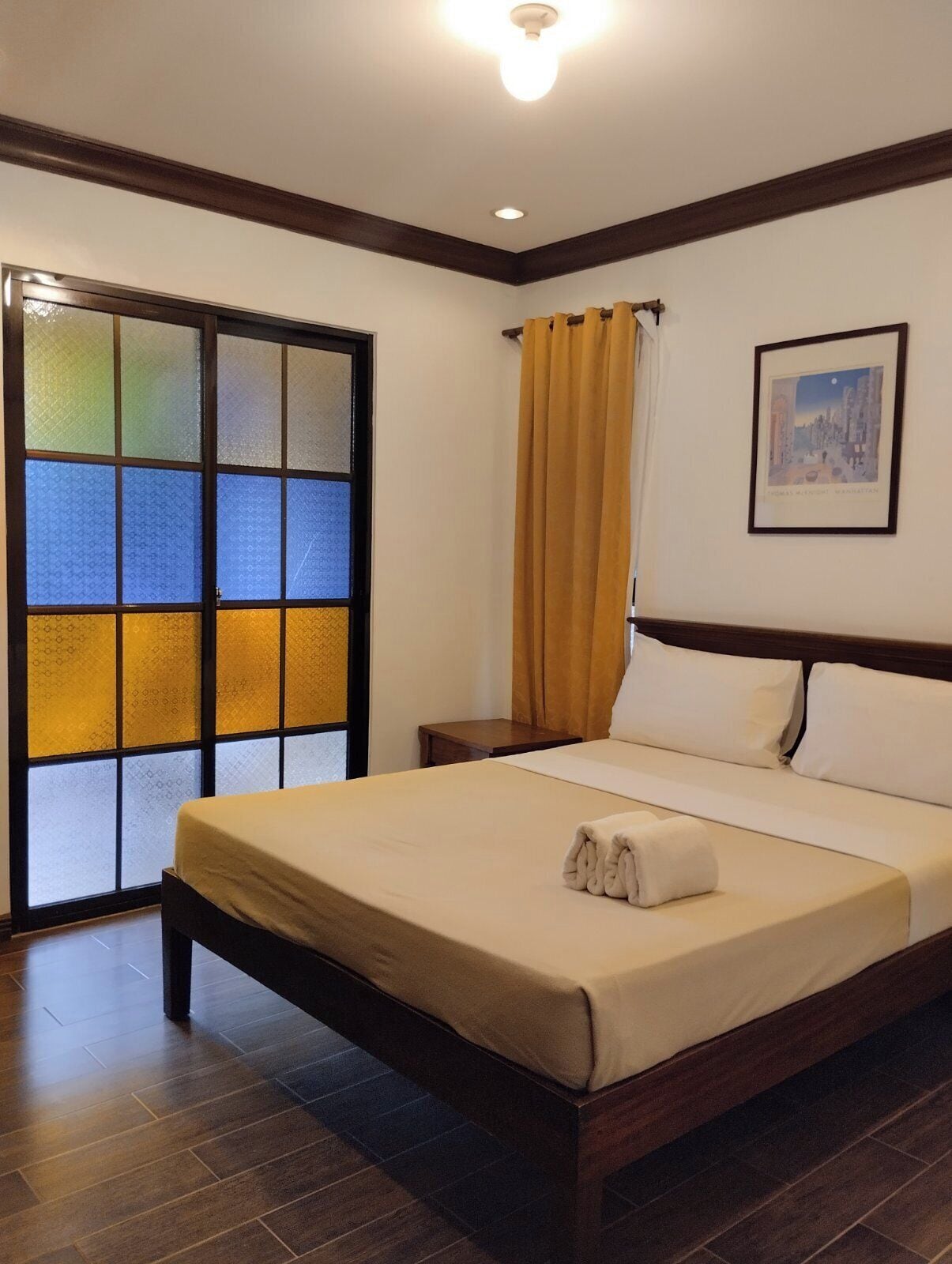 Una habitación de hotel con una cama, una mesita de noche, un cuadro en la pared y un ventanal corredero con paneles de vidrio esmerilado.