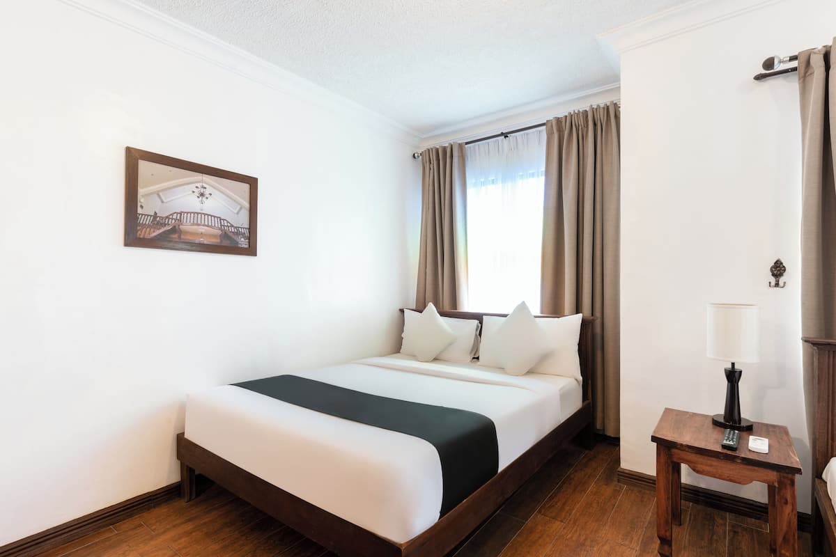 A hotel room with a large bed, a bedside table with a lamp, a framed picture on the wall, and a window with curtains.