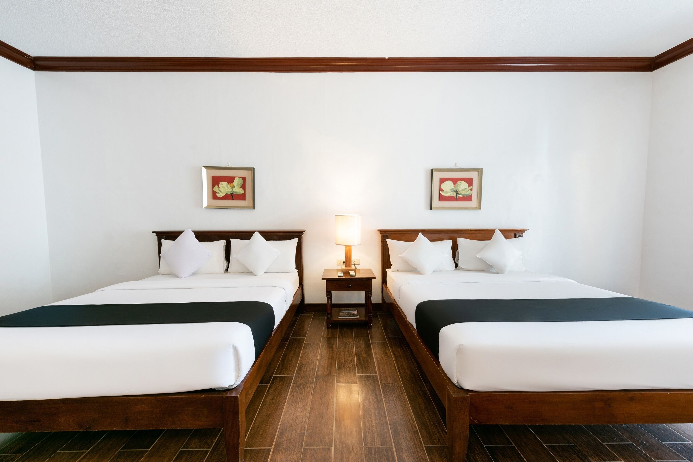 Ein Hotelzimmer mit zwei Einzelbetten, Holz-Kopfteilen, weißer Bettwäsche, einem kleinen Nachttisch mit Lampe sowie zwei gerahmten Blumenbildern an der Wand.