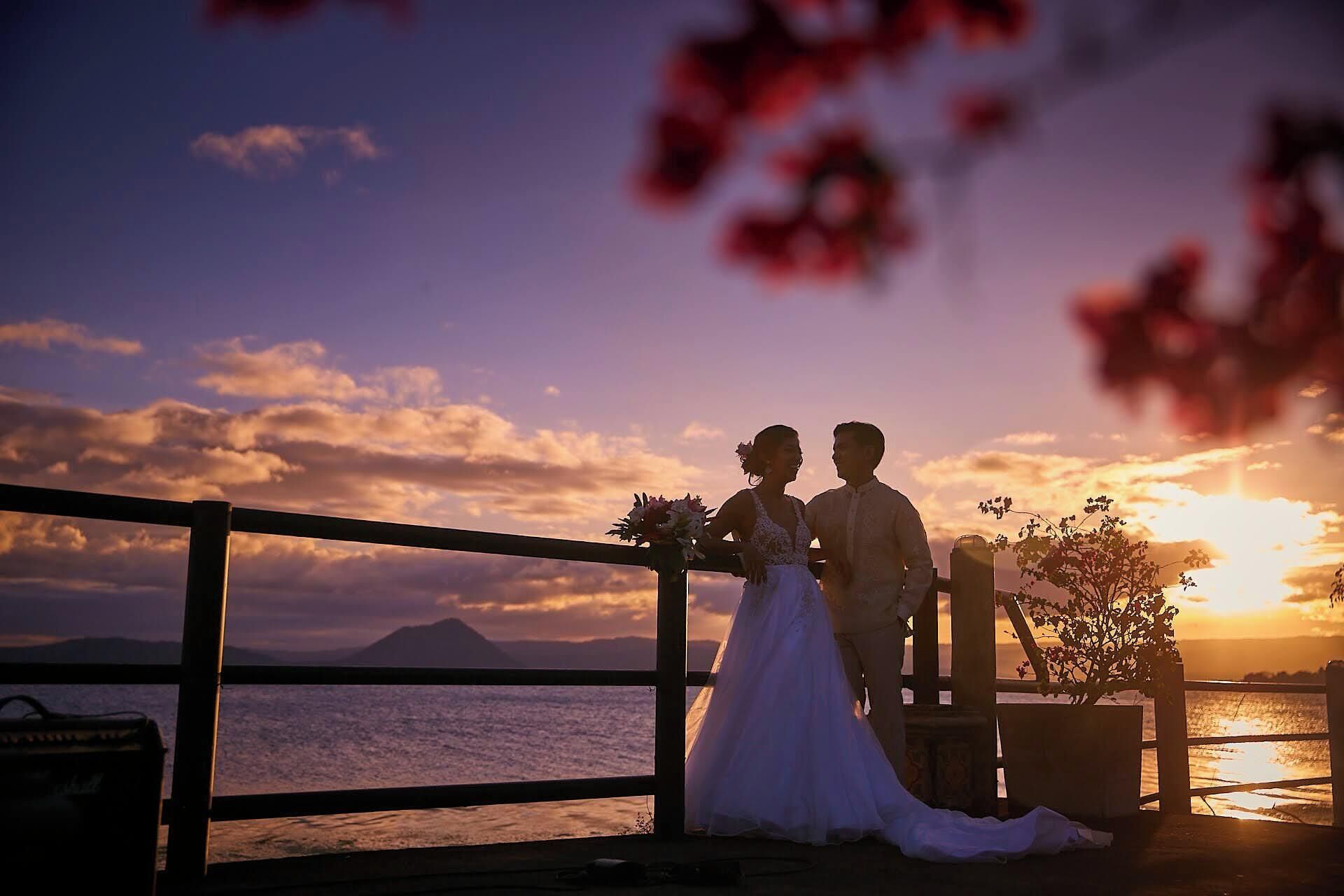 Una pareja de pie junto a una barandilla, con vista al atardecer sobre el agua.