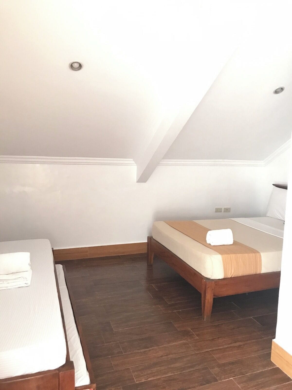 Una habitación pequeña y bien iluminada con piso de madera, una cama con colchón blanco y cabecera marrón, y una mesita con una toalla blanca doblada.