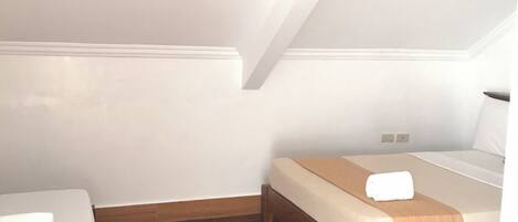 Una habitación pequeña y bien iluminada con piso de madera, una cama con colchón blanco y cabecera marrón, y una mesita con una toalla blanca doblada.