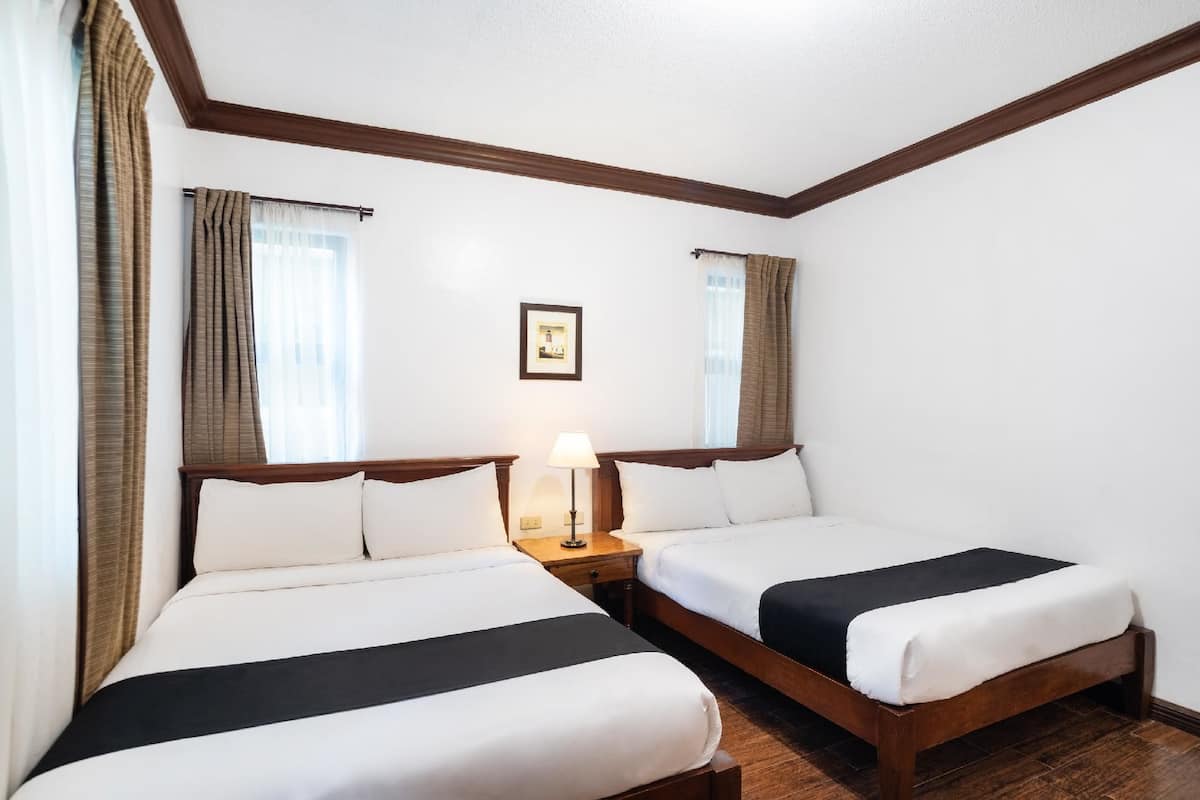 A hotel room with two beds, wooden headboards, white bedding, a nightstand with a lamp, and a framed picture on the wall.
