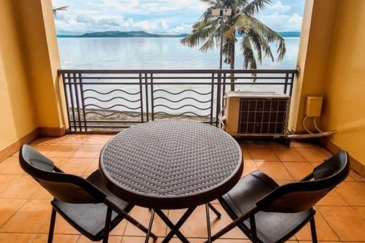A balcony with a round table and four chairs, overlooking a calm body of water and palm trees.