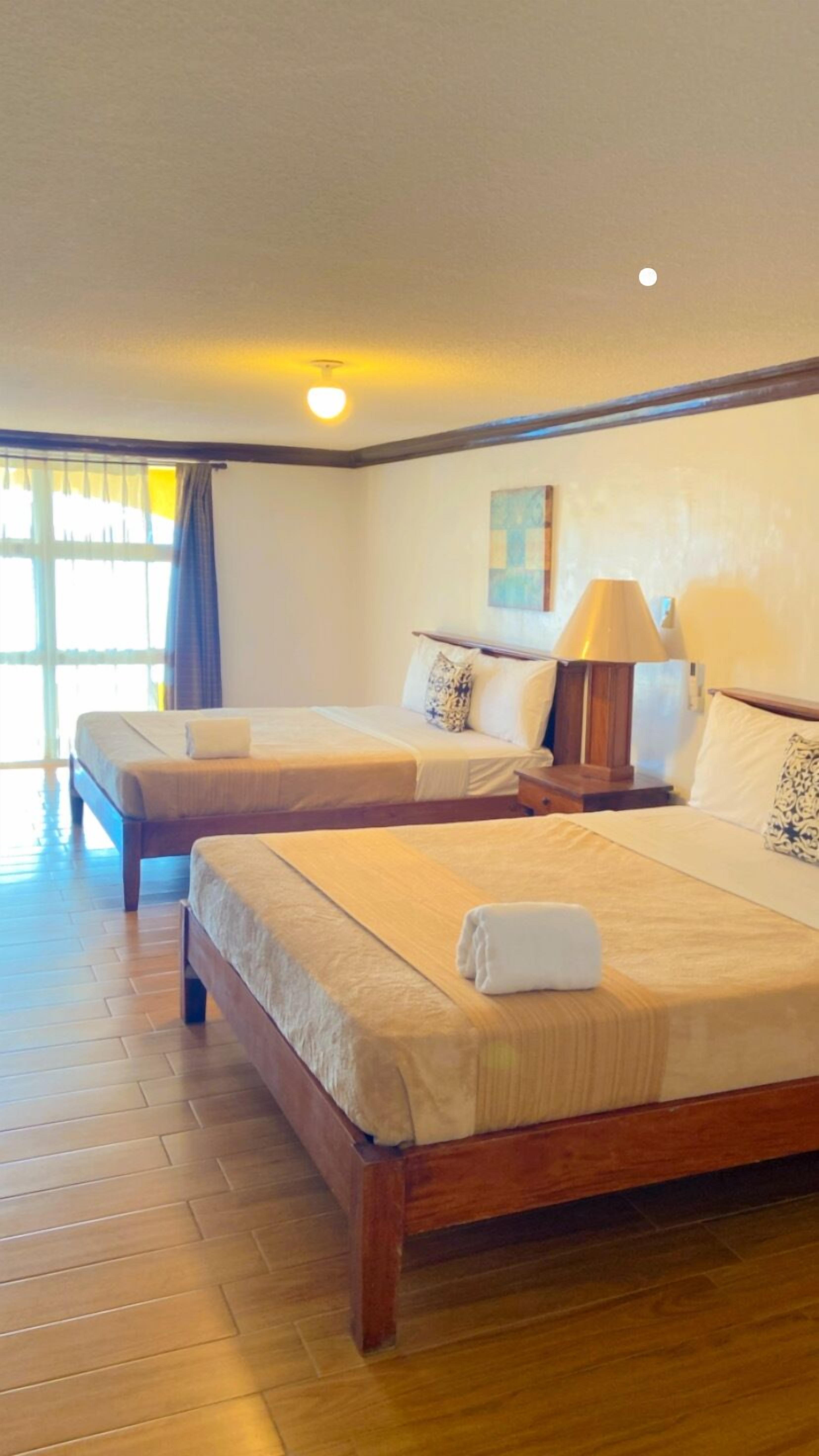 A hotel room with two wooden beds, a nightstand with a lamp, a painting on the wall, and a window with curtains.
