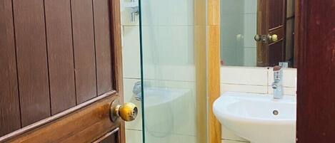 Un baño con puerta de madera, cabina de ducha con mampara de vidrio, lavabo blanco y espejo.