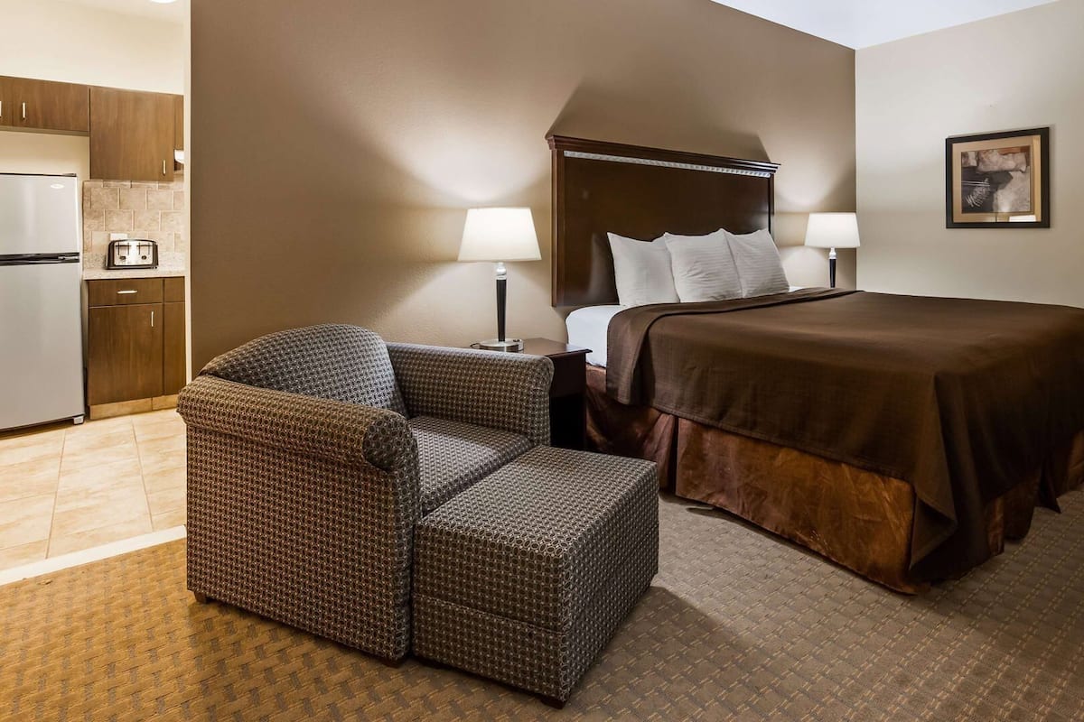 A hotel room with a bed, a chair, a nightstand, a lamp, a picture on the wall, and a kitchenette with a refrigerator.