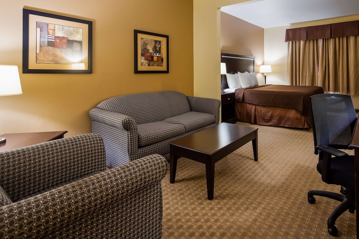 A hotel room with a patterned sofa, a small table, a desk with a chair, a bed with a brown cover, and two framed pictures on the wall.