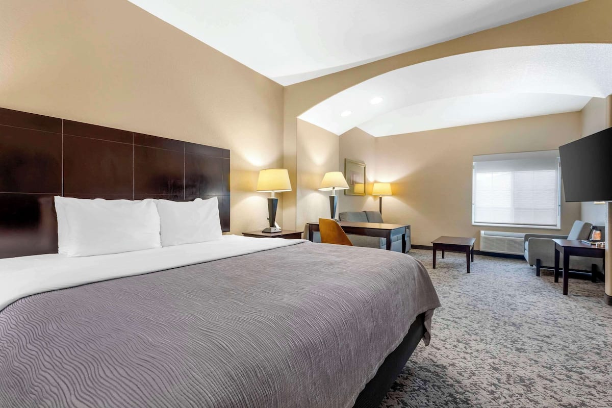 A hotel room with a large bed, two bedside tables with lamps, a desk with a chair, a TV, and a window with blinds.