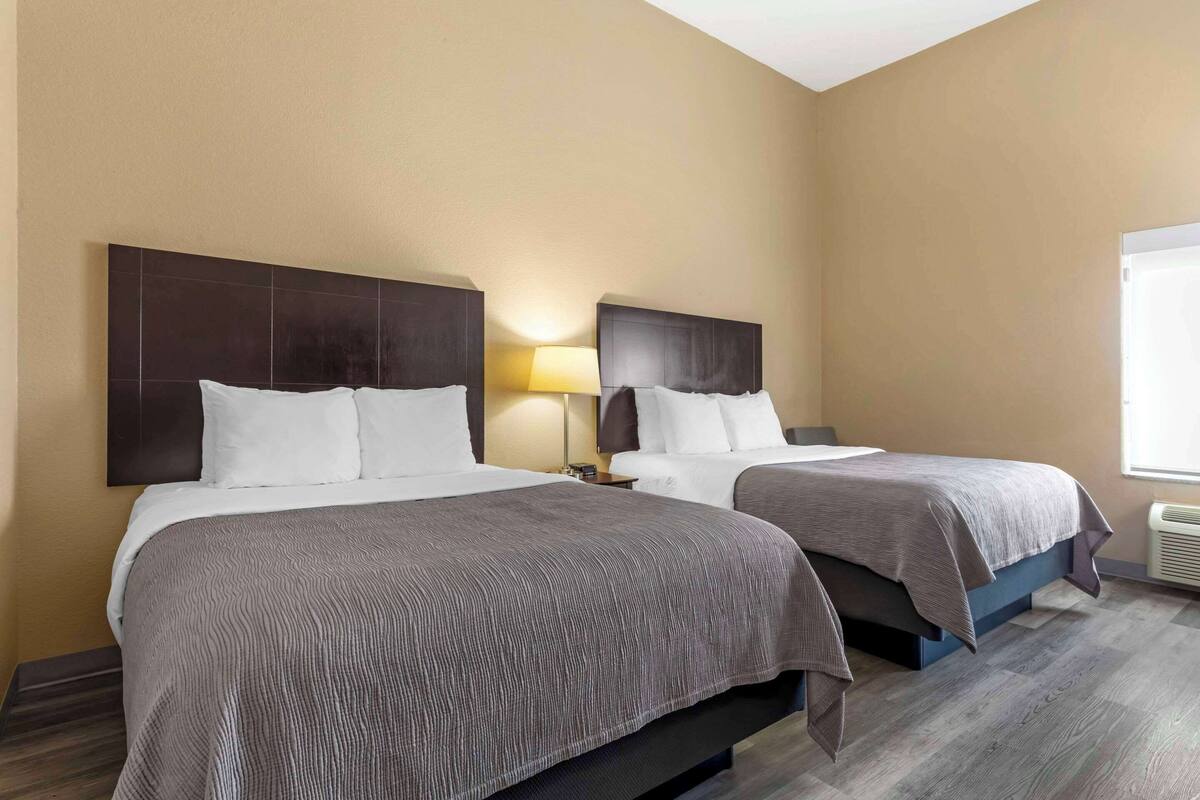 A hotel room with two beds, each with a dark headboard and white bedding. There is a nightstand with a lamp between the beds.