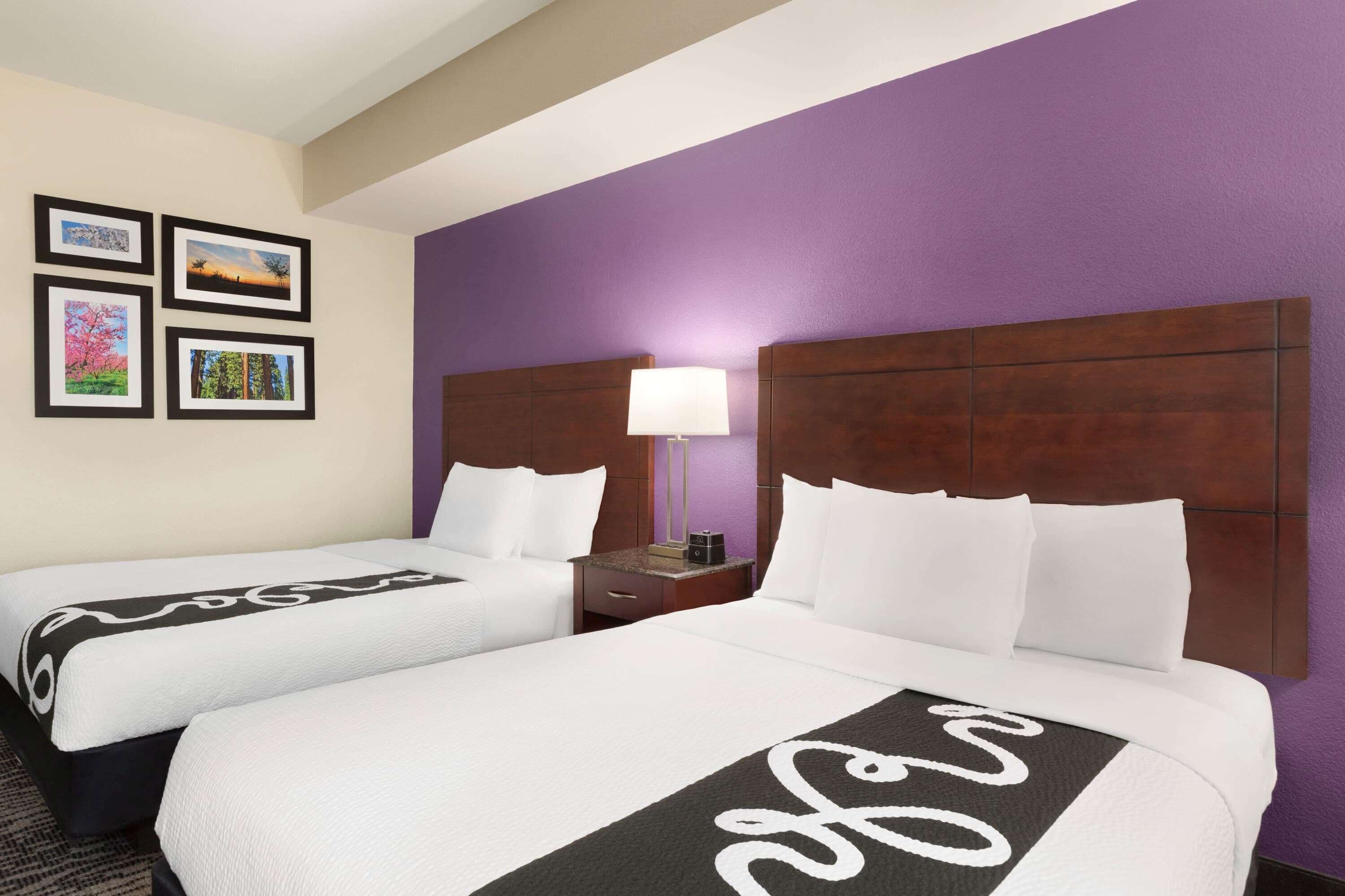 A hotel room with two beds, a nightstand, a lamp, and framed pictures on the wall.