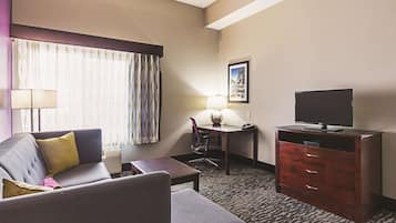 A hotel room with a sofa, a desk with a lamp, a television on a dresser, and a carpeted floor.
