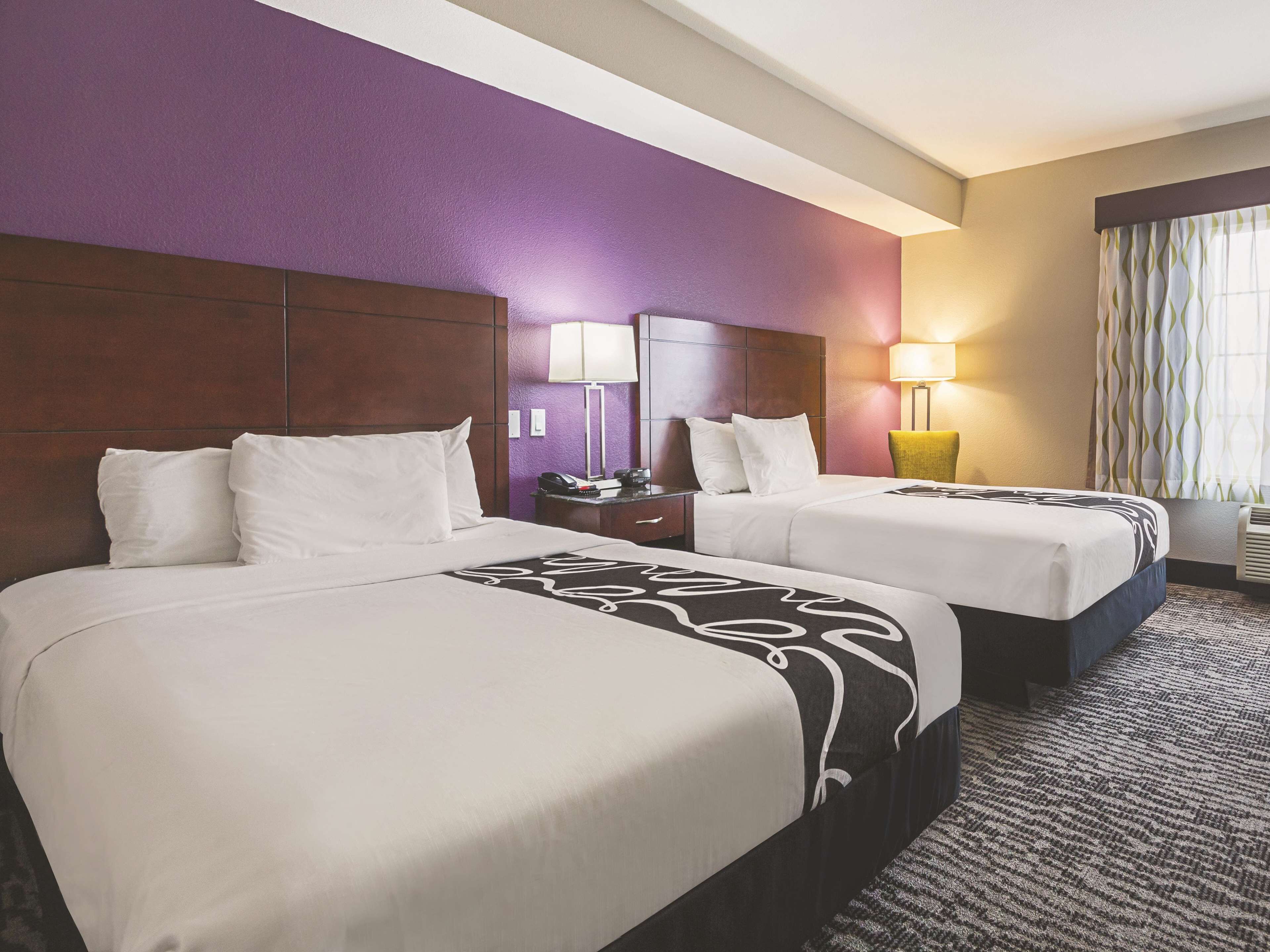 A hotel room with two beds, purple walls, a wooden headboard, a nightstand with a lamp, and a window with curtains.