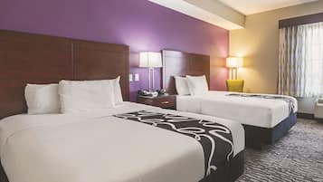 A hotel room with two beds, purple walls, a wooden headboard, a nightstand with a lamp, and a window with curtains.