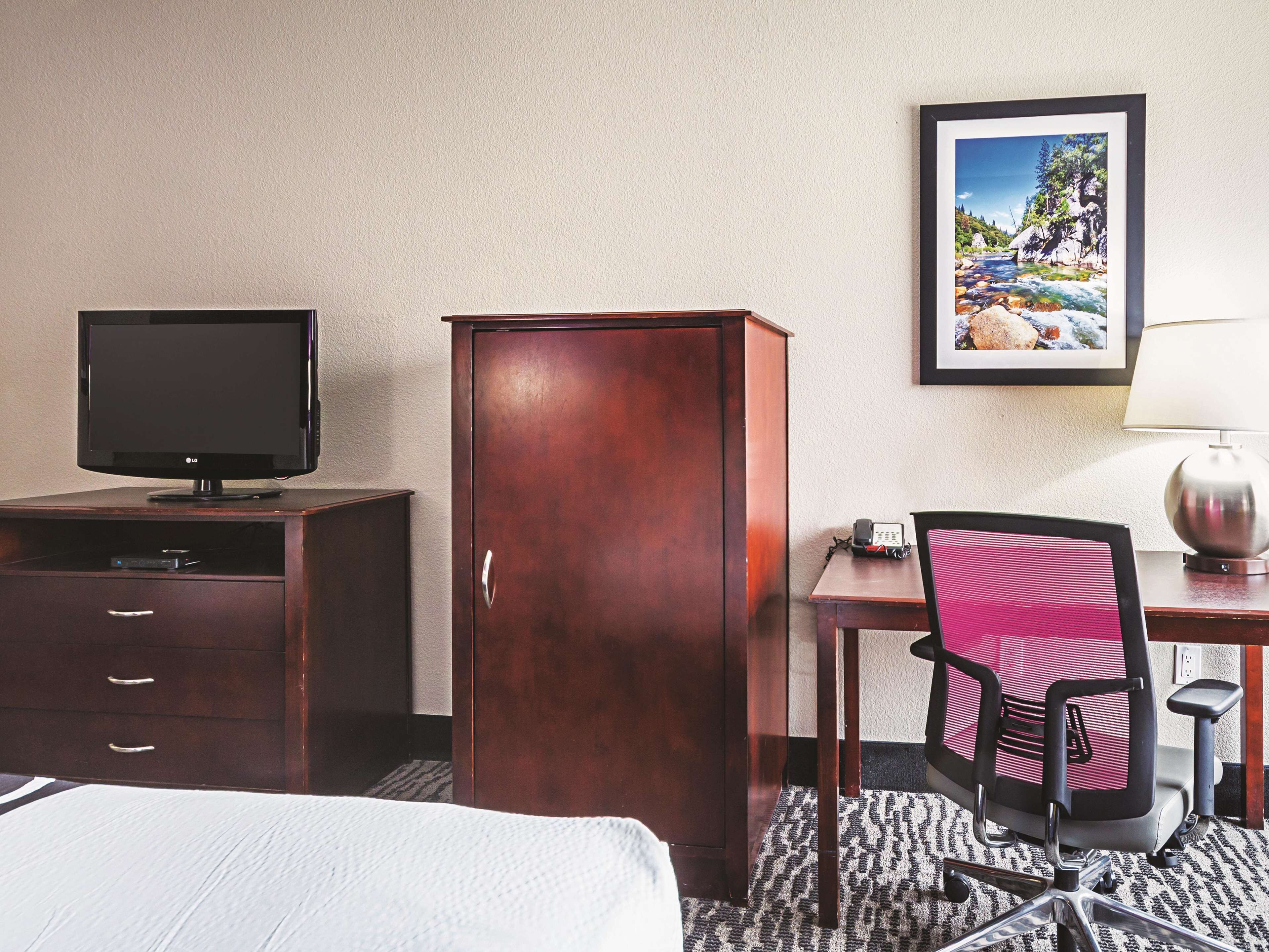 Una habitación de hotel con una cama, un escritorio con silla, un televisor, un armario y un cuadro en la pared.