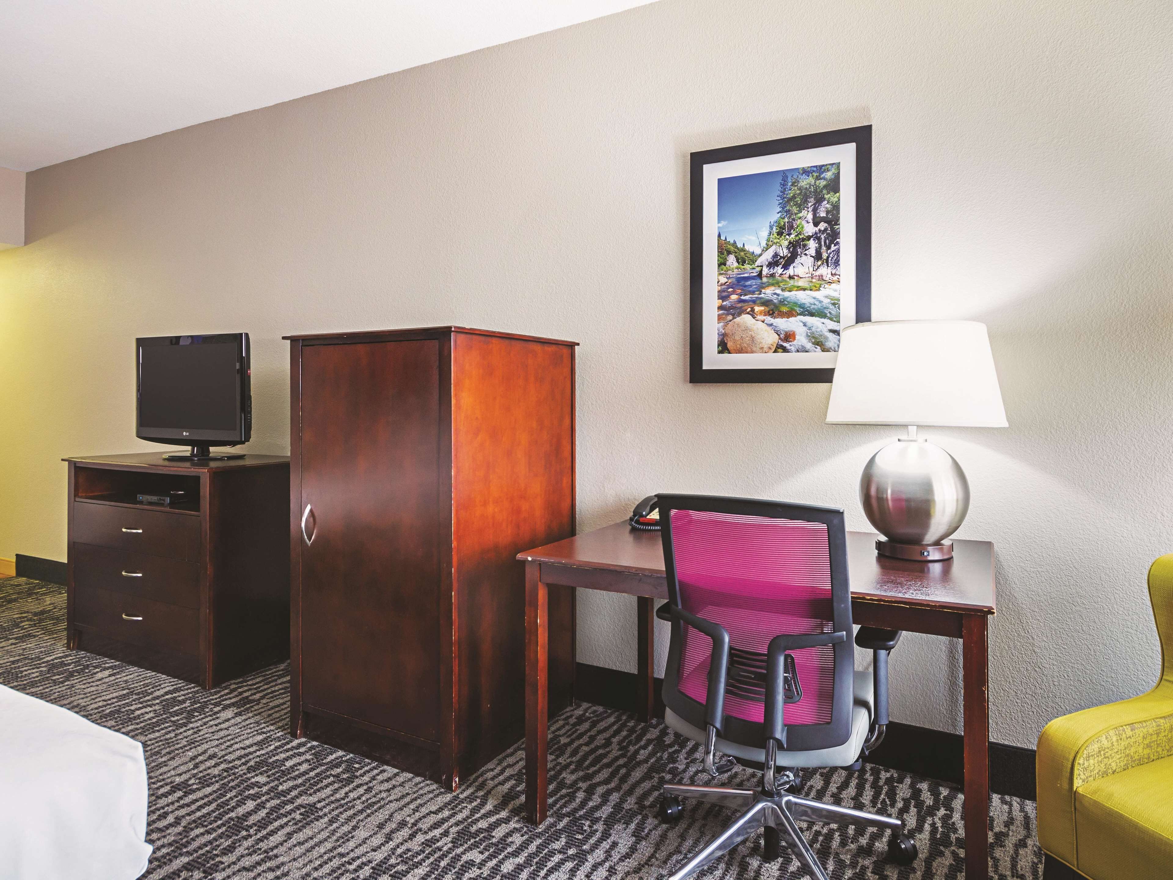 A hotel room with a desk, chair, TV, wardrobe, lamp, and a framed picture on the wall.