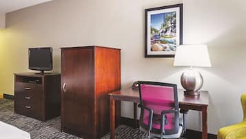 A hotel room with a desk, chair, TV, wardrobe, lamp, and a framed picture on the wall.