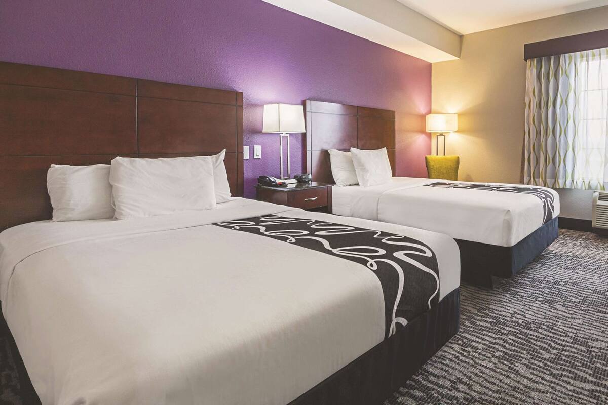 Une chambre d’hôtel avec deux lits, des murs violets, une tête de lit en bois, une table de chevet, une lampe et une fenêtre avec des rideaux.