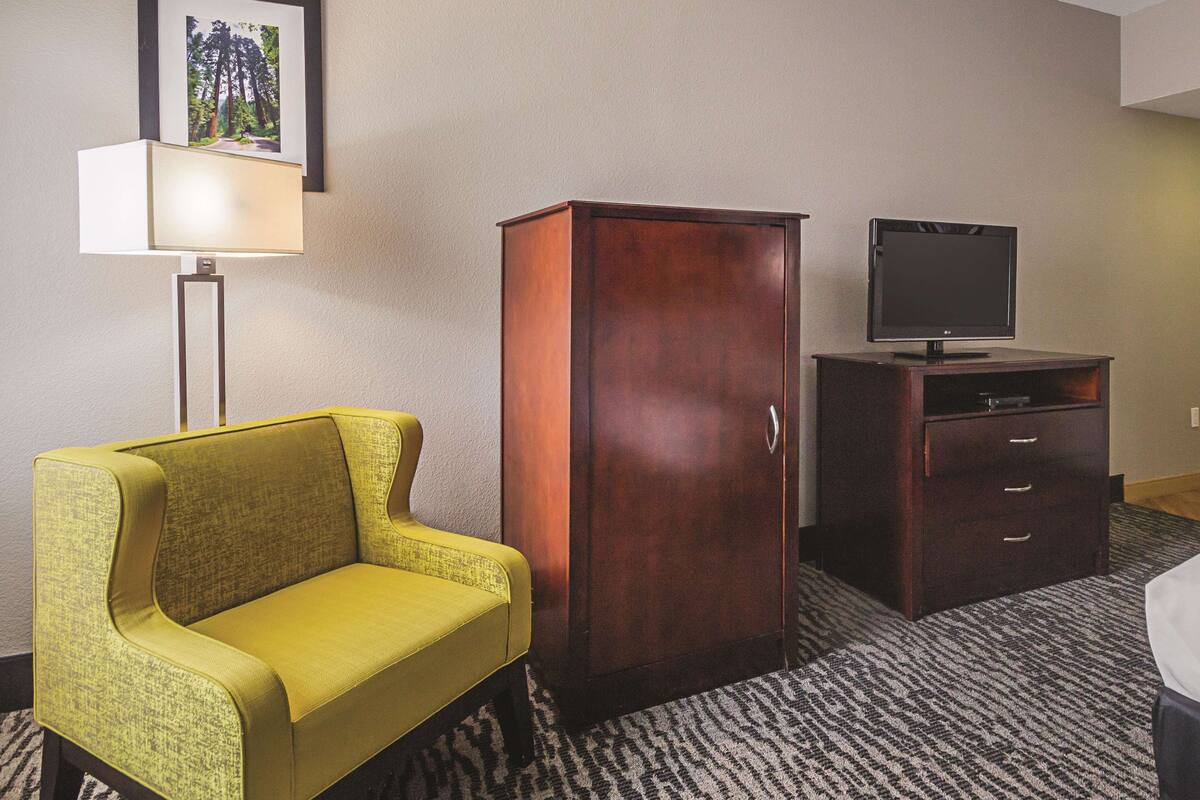 Une chambre d’hôtel avec un fauteuil jaune, une armoire en bois, un téléviseur posé sur un meuble et un tableau encadré accroché au mur.