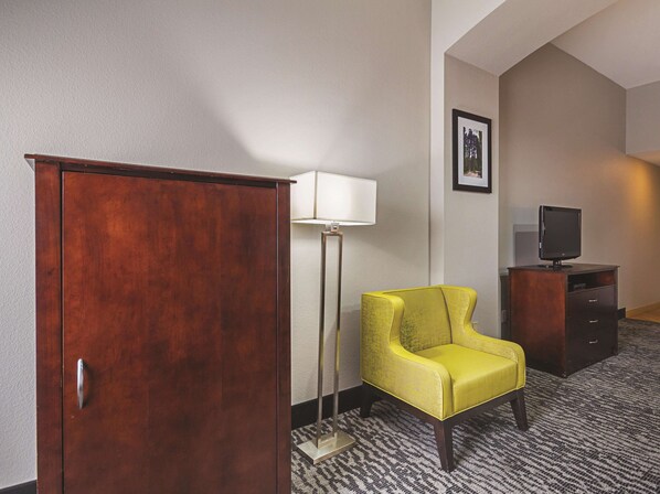 A hotel room with a wooden wardrobe, a yellow armchair, a floor lamp, a television on a wooden cabinet, and a carpeted floor.